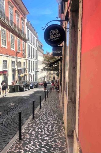 Photo of round sign of Dos Ovos Moles at the entrance with pedestrians walking on a tiled sidewalk and on a cobblestone street in front of it
