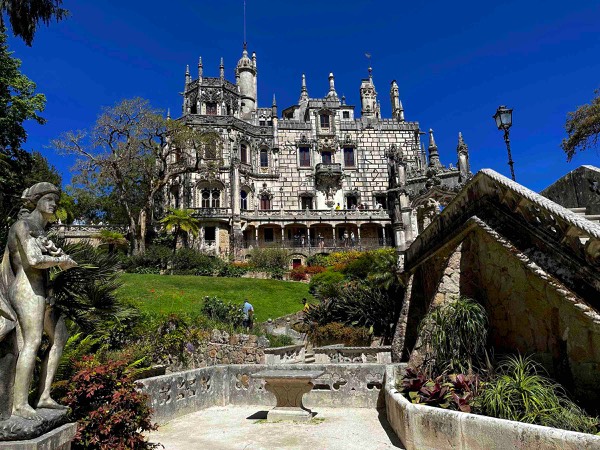 Photo of a palace on top of a hilly ground with a garden below it, garden statues to the left and stone stairway to the right